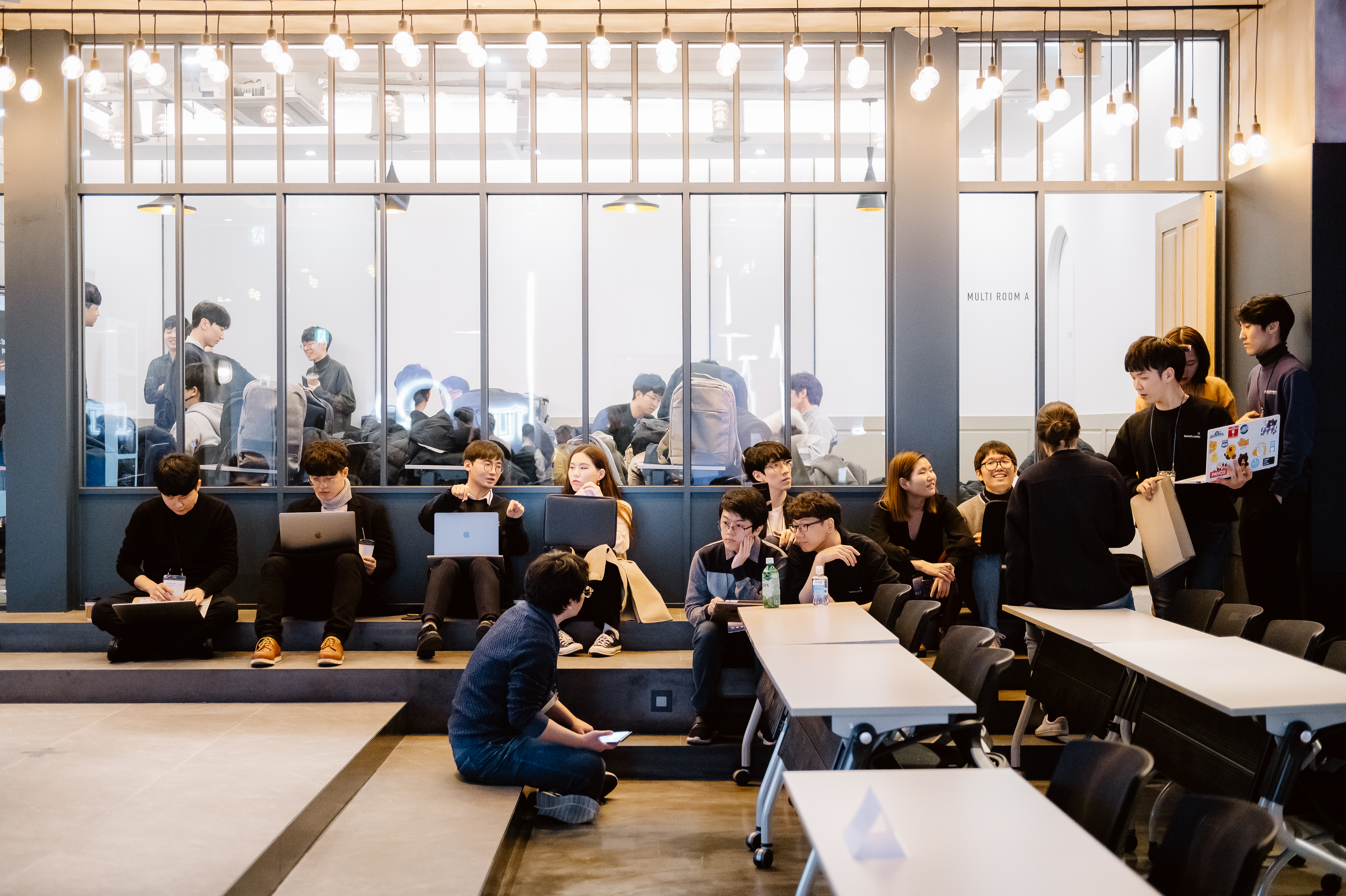 다양한 교육생들이 계단식 공간과 테이블에 자유롭게 앉아 노트북을 사용하거나 대화를 나누며 함께 학습하는 모습의 실습 사진입니다.